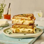 Stacked grilled cheese sandwich on a white plate garnished with herbs, surrounded by cheese slices, fresh herbs, and glasses of beverages on a light table.