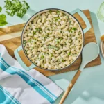 A light blue pot filled with creamy pasta salad topped with peas and herbs, surrounded by fresh basil, parsley, toasted bread slices, a glass of water, and a striped kitchen towel on a pale blue surface.