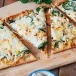 Sliced white pizza with melted cheese, spinach, and herbs on a wooden board, with sauce bowls and garnish visible.
