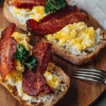 Three open-faced breakfast sandwiches on toasted bread with scrambled eggs, crispy bacon strips, and fresh parsley garnish on a wooden board.