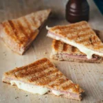 Three triangular sandwiches with cinnamon-sugar coating and melted cheese filling on a wooden surface with a dark bottle nearby.