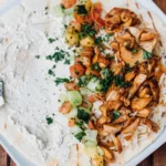 White plate with creamy dip, crispy fried onions, diced cucumbers, and fresh parsley garnish on wooden table.