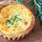 Savory tart with golden custard filling and chopped chives, garnished with fresh rosemary on wooden surface.