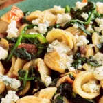 Pasta salad with orecchiette, sautéed greens, crumbled cheese, roasted red peppers, and black pepper on a green plate.