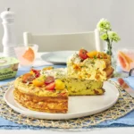 Elegant brunch spread featuring herb-topped omelette with tomatoes on white plate, pink beverages, garlic alouette spread, fresh flowers, and table setting with blue linens.