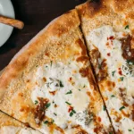 Freshly baked pizza with melted cheese, herbs, and red pepper flakes on parchment paper, with a wooden spoon and chili seasoning bowl nearby.