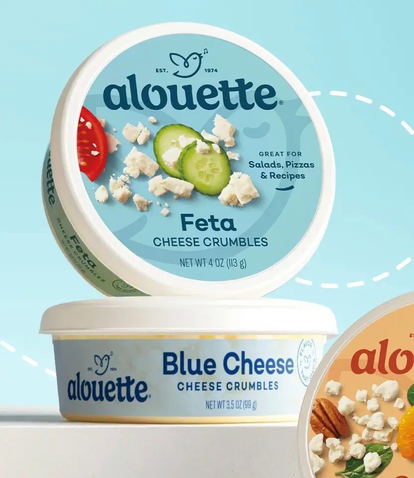 Two Alouette cheese crumble containers displayed against a light blue background: Feta (top, 4 oz) and Blue Cheese (bottom, 3.5 oz), with fresh vegetables and ingredients shown on packaging.