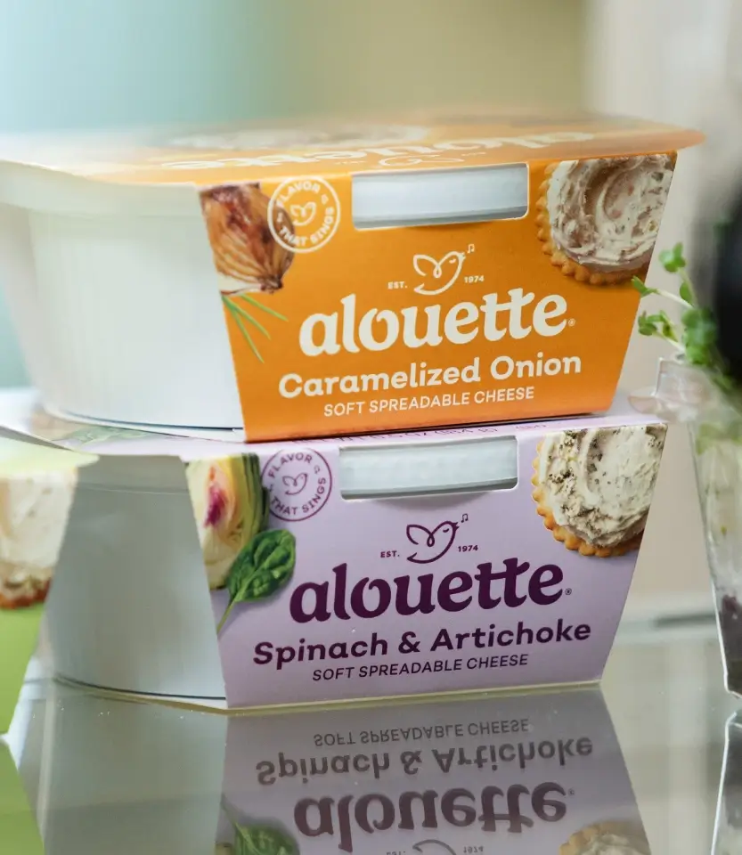 Two stacked Alouette soft spreadable cheese containers: Caramelized Onion (orange) on top and Spinach & Artichoke (purple) below, displayed on a reflective surface.