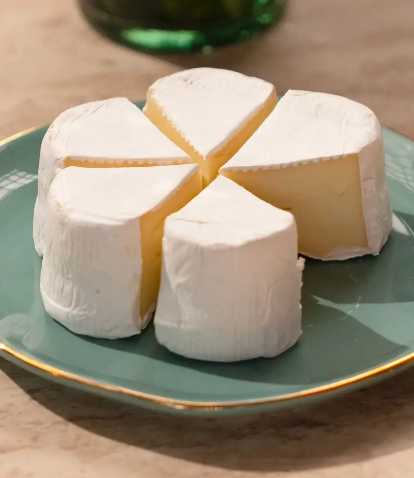 Sliced cheese wheel with white rind tied with golden ribbon on a sage green plate with gold rim.