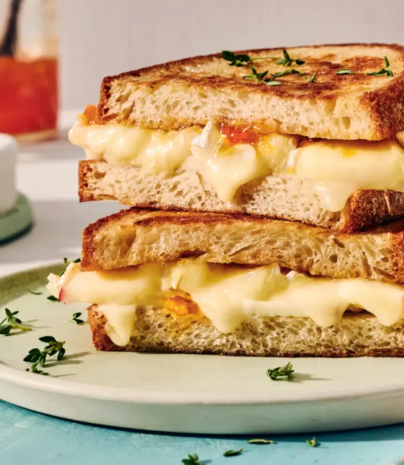 Grilled cheese sandwich stacked and cut diagonally on white plate, garnished with fresh thyme, with beverage in background.