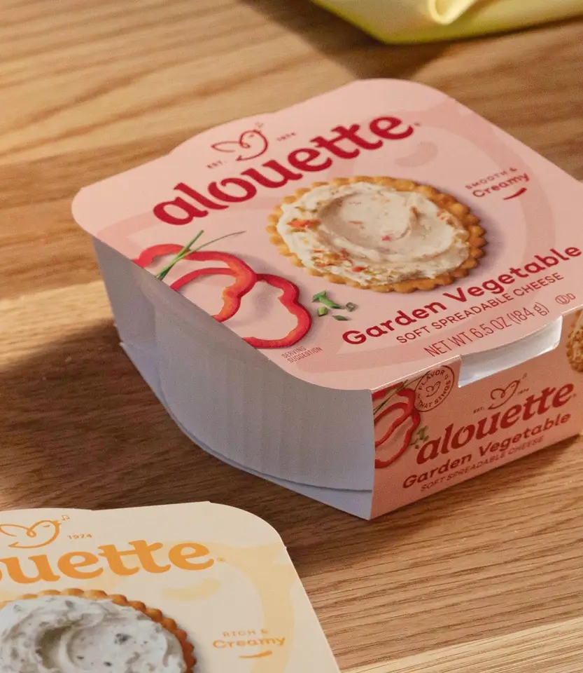 Two Alouette Garden Vegetable soft spreadable cheese containers on a wooden surface, with crackers visible in the background.