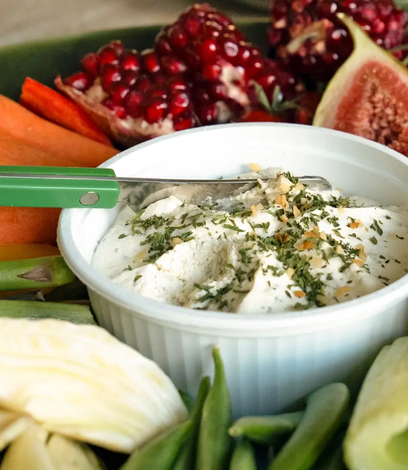 White bowl of herb-topped dip surrounded by fresh vegetables including carrots, green beans, pomegranate, and figs, with a green serving spoon.