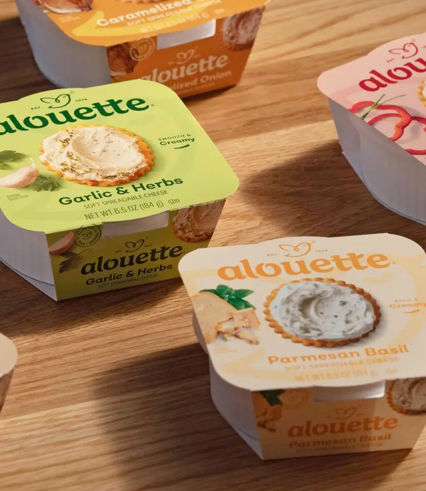 Multiple Alouette cheese spread containers in various flavors including Garlic & Herbs and Parmesan Basil arranged on a wooden surface.