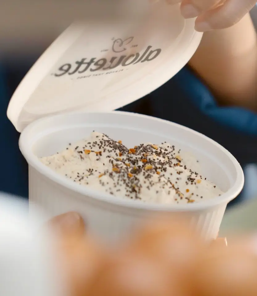 Person holding an open white container with creamy soup or dip topped with herbs and spices.