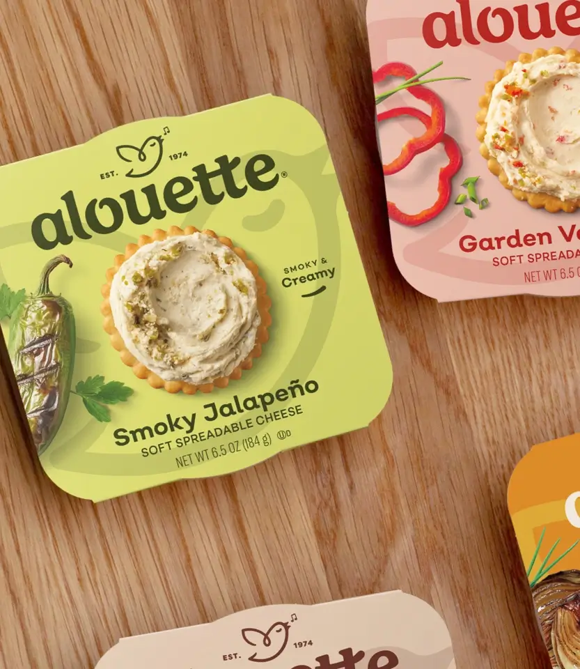 Alouette soft spreadable cheese packages displayed on wood surface, featuring Smoky Jalapeño and Garden Vegetable flavors with crackers topped with cheese spread.