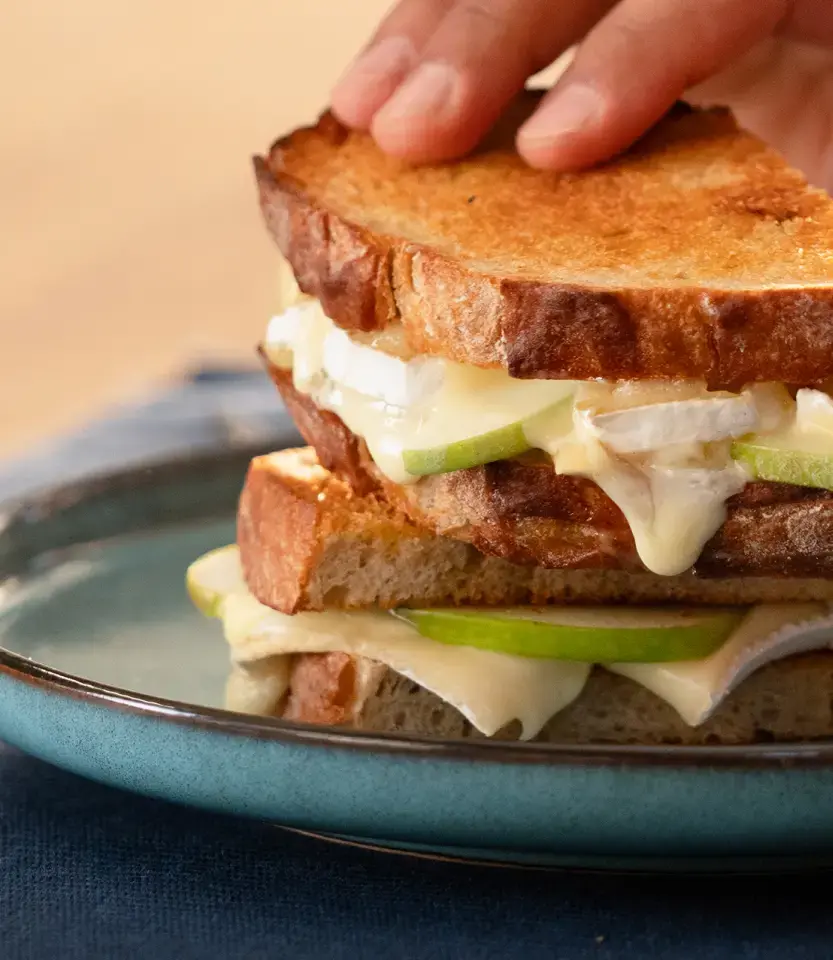 Hand holding a grilled cheese and apple sandwich with bacon on toasted bread, served on a blue plate.