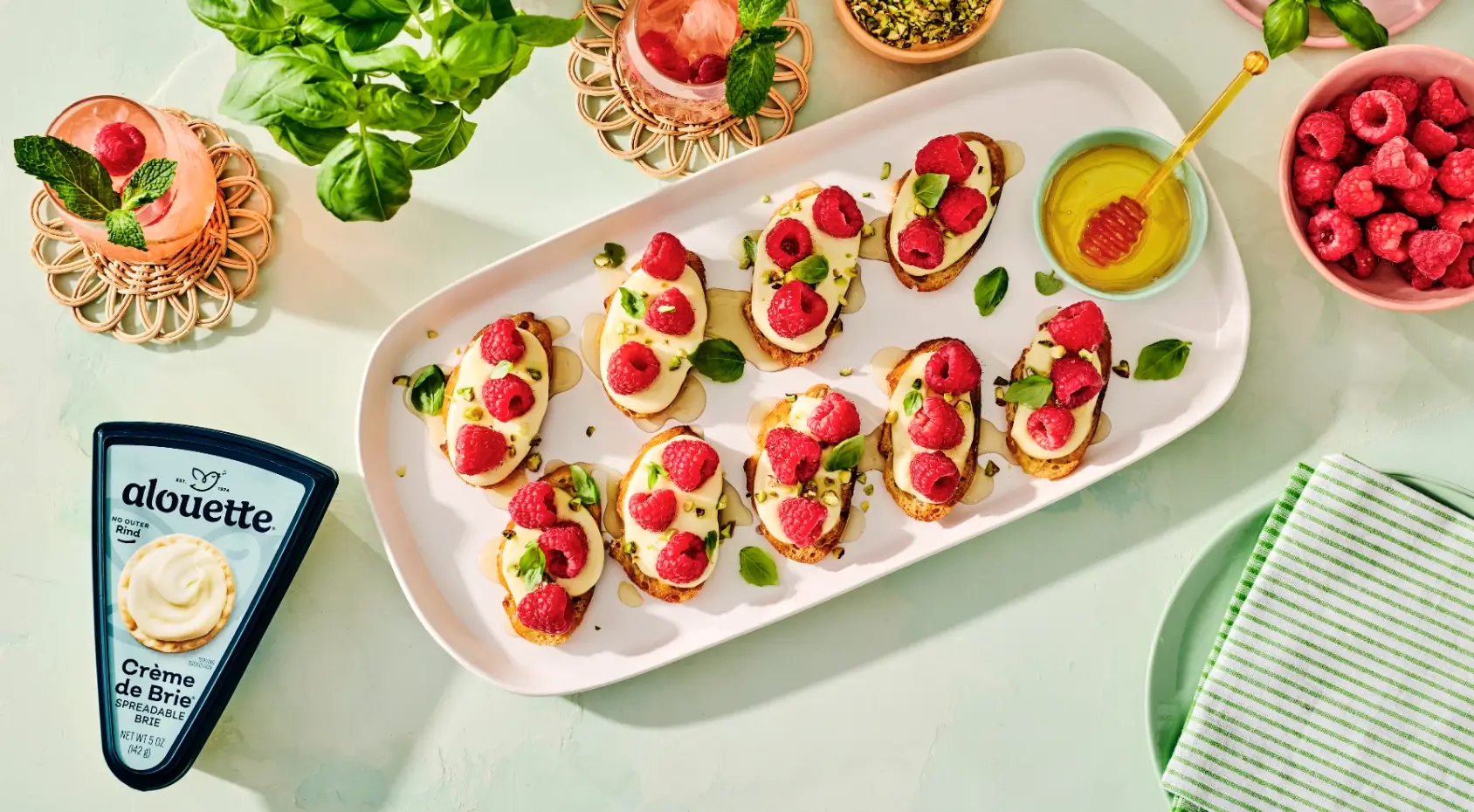 Elegant appetizer spread with raspberry crostini on creamy cheese base, fresh raspberries, mint garnish, cocktails, and Alouette Crème de Brie on white surface.