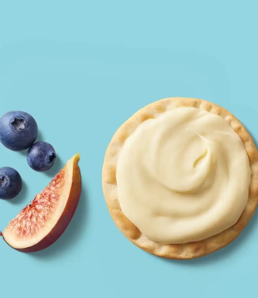 Blueberries and a fig slice beside a cream-topped cracker on a light blue background.