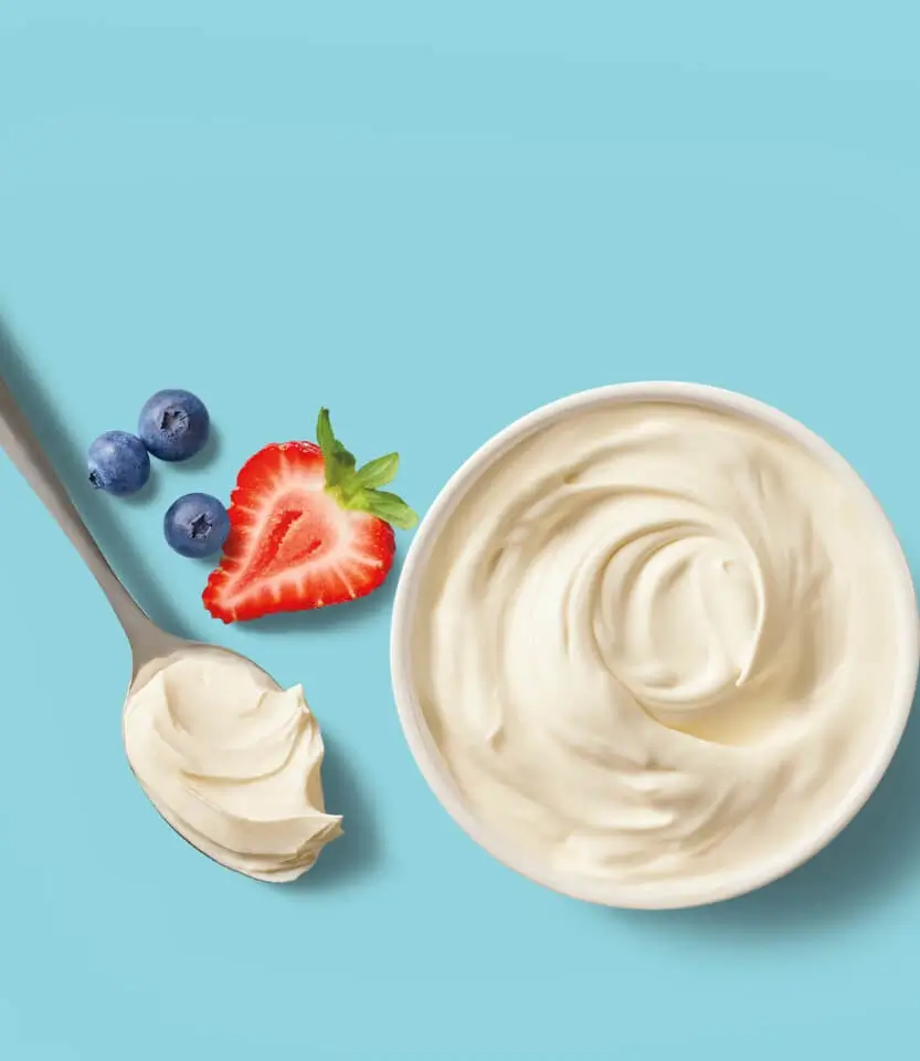 Creamy yogurt in a bowl with a spoon, fresh blueberries, and sliced strawberry on a light blue background.