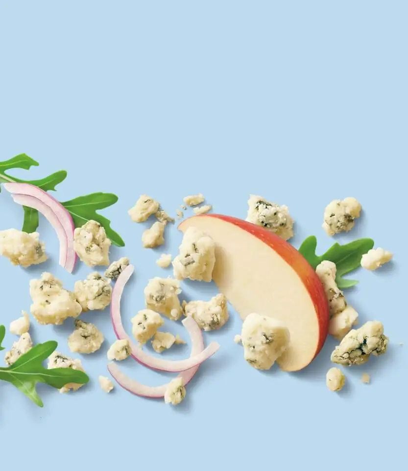 Apple slice with red skin surrounded by cauliflower florets, pink onion slices, and green herbs on blue background.