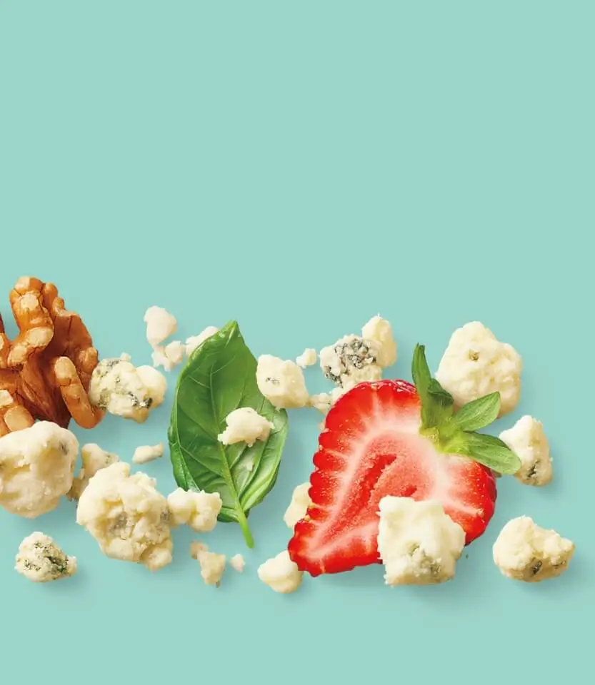 Sliced strawberry surrounded by popcorn, walnuts, basil leaves, and blue cheese crumbles on a turquoise background.