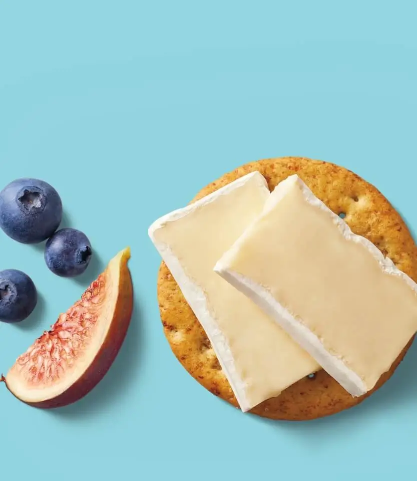 Toasted bagel with cream cheese slices, fresh blueberries, and a fig wedge on a light blue background.