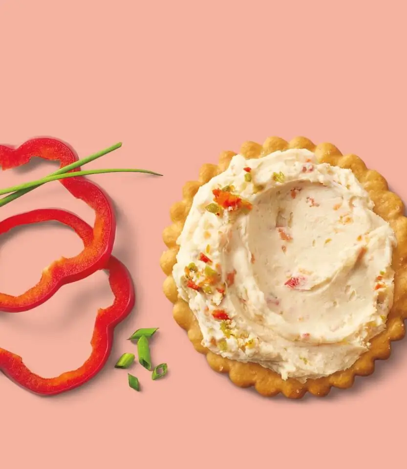 Decorative cookie with cream frosting topped with red pepper pieces and green garnish, alongside sliced red bell pepper on pink background.