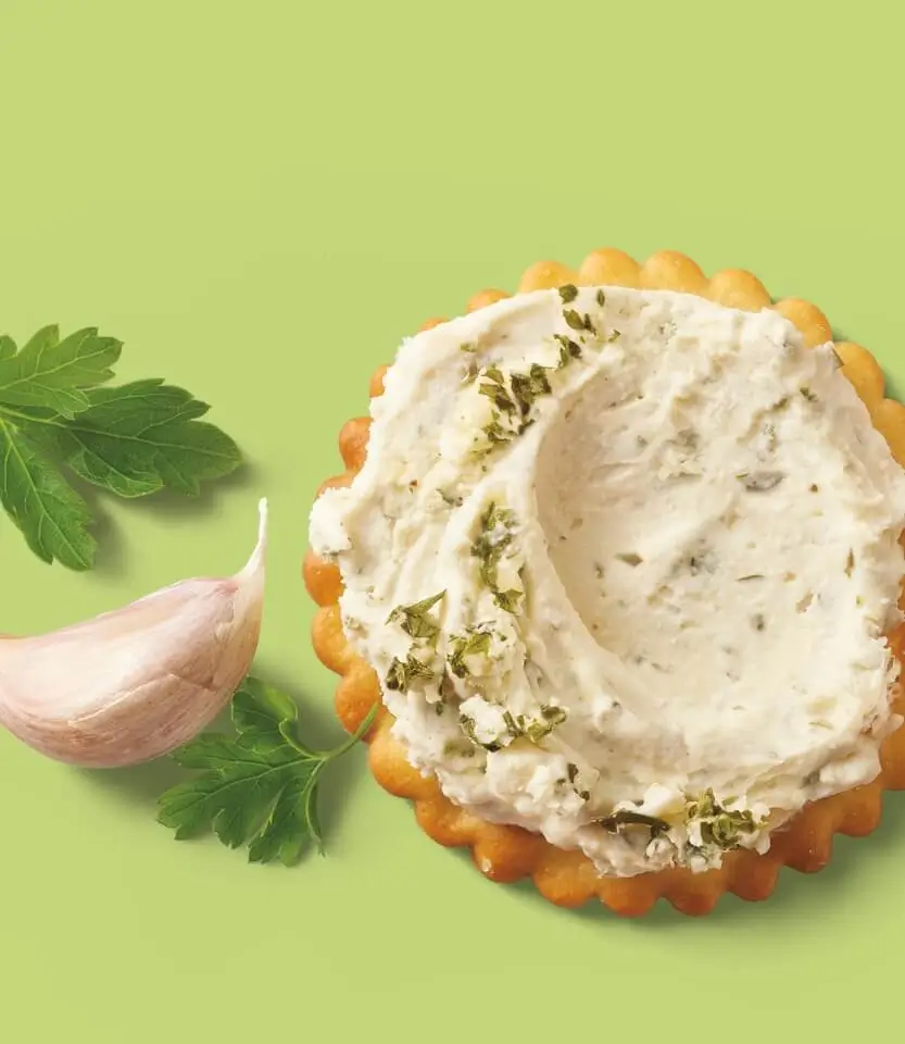 Herb-topped cream cheese spread on a decorative cracker with fresh garlic clove and parsley on a green background.