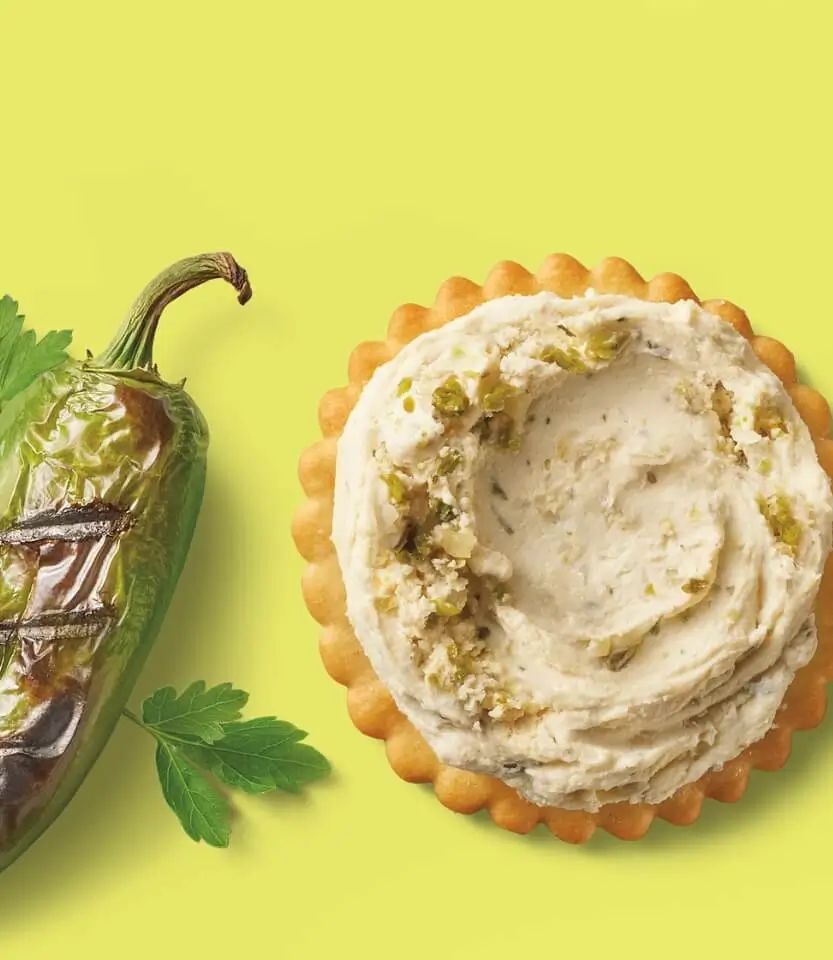 Roasted poblano pepper beside a scalloped-edge tart topped with cream cheese frosting and crushed pistachios on yellow background.