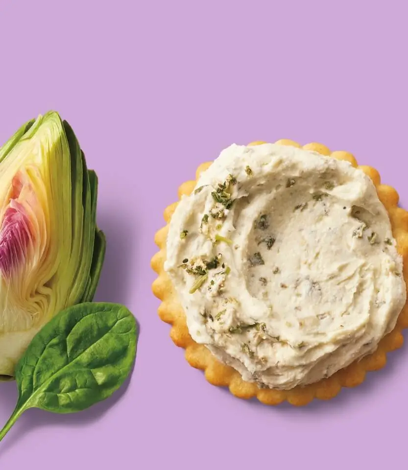 A cream-topped cracker with herb garnish next to a sliced artichoke and spinach leaf on a purple background.