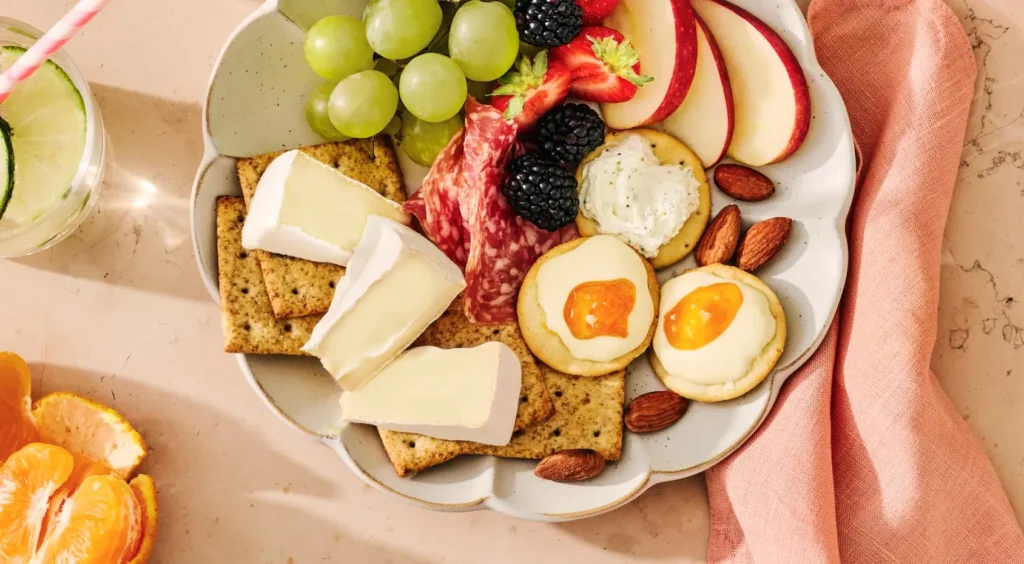 Charcuterie board with cheese wedges, crackers, cured meats, almonds, grapes, strawberries, blackberries, apples, and jam-topped crackers on a beige plate.
