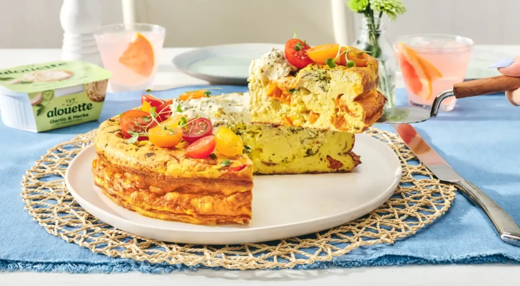 A plated omelet with herbs and diced peppers sits on a white plate with a woven placemat, accompanied by pink beverages and fresh flowers at a blue-clothed table setting.