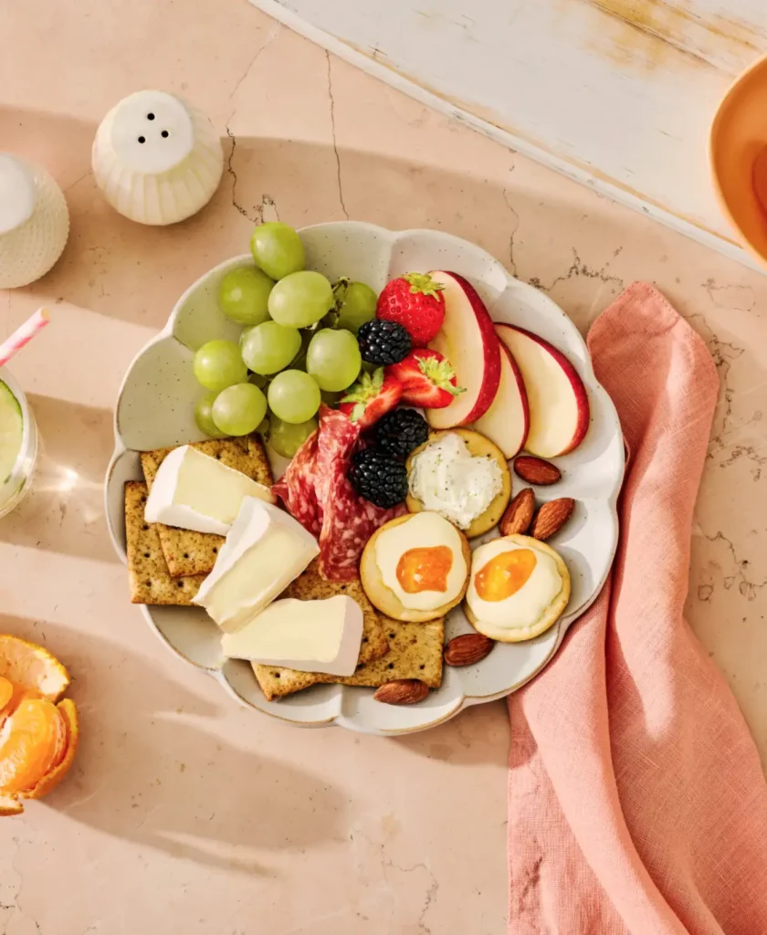 A decorative charcuterie board with green grapes, strawberries, blackberries, apple slices, cured meats, cheese, crackers, almonds, and apricot-topped crostini on a marble surface.