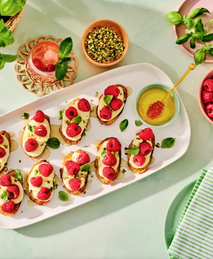 Elegant open-faced appetizers with cream cheese, fresh raspberries, and pistachios on toasted bread, arranged on a white plate with mint garnish and honey drizzle.