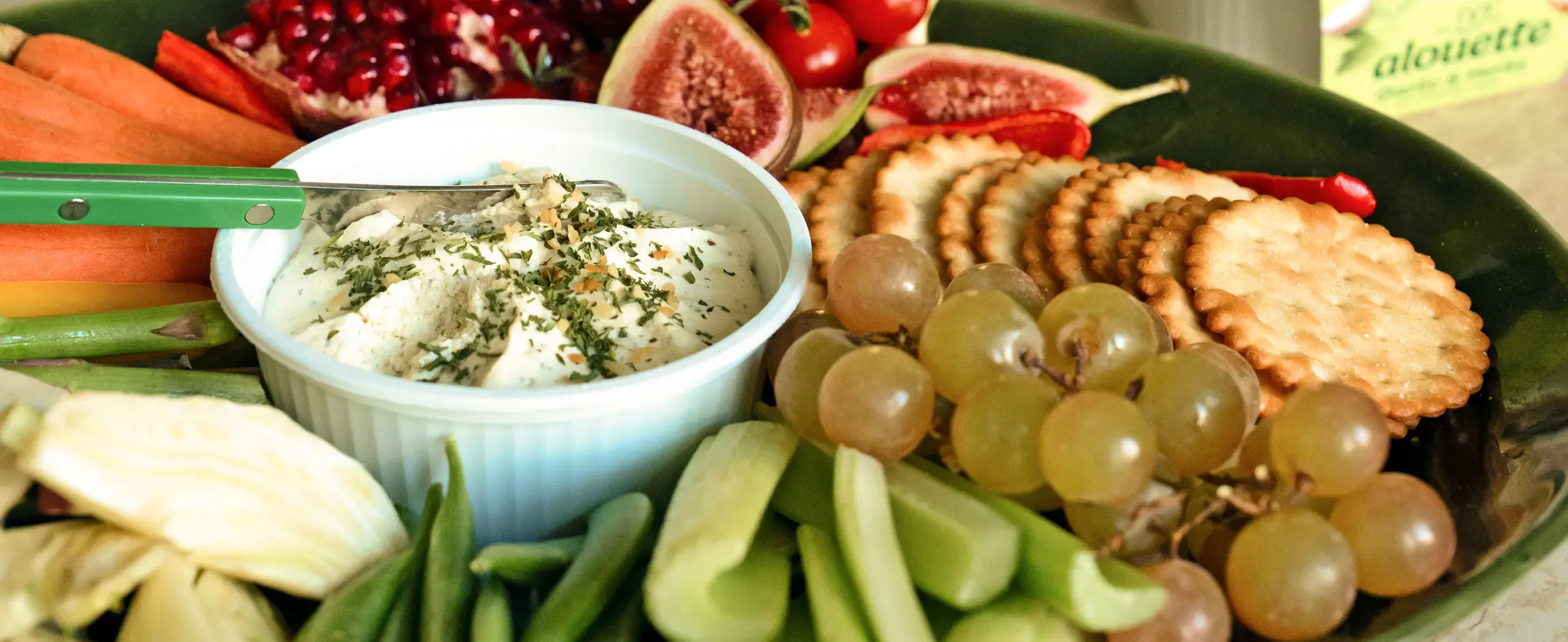 Charcuterie board with herb dip, fresh vegetables, grapes, crackers, figs, and pomegranate on green tray.
